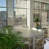 An office space with two tables each surrounded by chairs, two plants and a scenic view through the window with blinds.